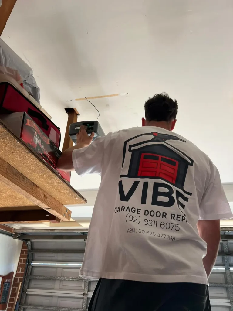 Technician repairing a garage door opener, wearing a branded Vibe Garage Door Repair shirt.
