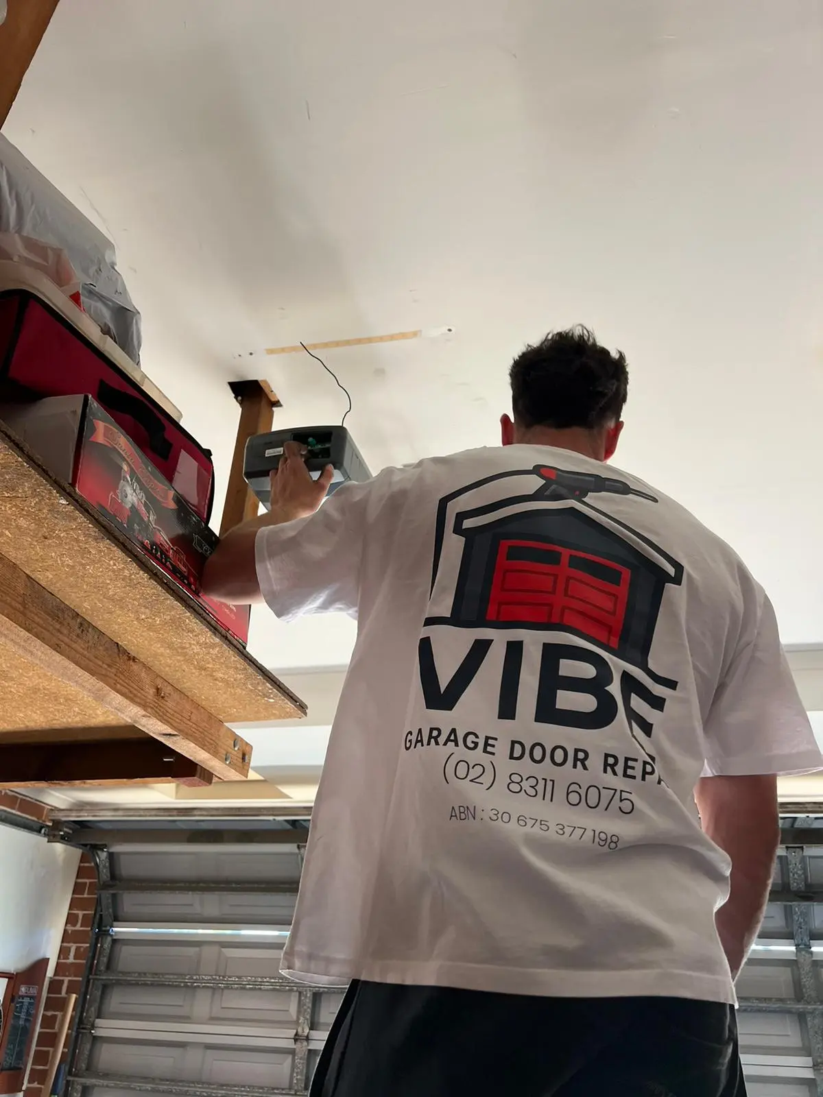 Technician repairing a garage door opener, wearing a branded Vibe Garage Door Repair shirt.